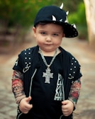 A young child is dressed in a punk-inspired outfit, featuring a black cap adorned with metallic spikes, a necklace with a large cross pendant, and a black shirt with studded details. The child's arms are covered in tattoo-like sleeves.