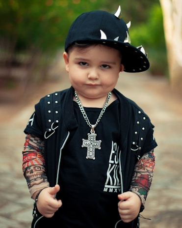 A young child is dressed in a punk-inspired outfit, featuring a black cap adorned with metallic spikes, a necklace with a large cross pendant, and a black shirt with studded details. The child's arms are covered in tattoo-like sleeves.