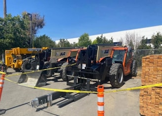Several heavy construction vehicles are lined up in an outdoor area. They are behind a barrier of yellow caution tape and orange traffic cones. The ground is paved and there are trees in the background, with a building partially visible. Stacks of wooden boards are on one side.