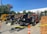 Several heavy construction vehicles are lined up in an outdoor area. They are behind a barrier of yellow caution tape and orange traffic cones. The ground is paved and there are trees in the background, with a building partially visible. Stacks of wooden boards are on one side.
