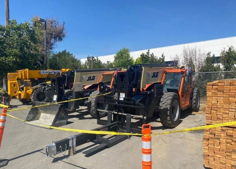 Several heavy construction vehicles are lined up in an outdoor area. They are behind a barrier of yellow caution tape and orange traffic cones. The ground is paved and there are trees in the background, with a building partially visible. Stacks of wooden boards are on one side.
