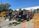 Several heavy construction vehicles are lined up in an outdoor area. They are behind a barrier of yellow caution tape and orange traffic cones. The ground is paved and there are trees in the background, with a building partially visible. Stacks of wooden boards are on one side.