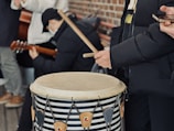 A group of people is gathered for a musical performance, with one individual playing a large drum using drumsticks. Another person is seen with a guitar, while another holds a smartphone. Everyone is wearing warm clothing, indicating a cold environment. The setting is casual and outdoors, with a brick wall visible in the background.
