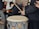 A group of people is gathered for a musical performance, with one individual playing a large drum using drumsticks. Another person is seen with a guitar, while another holds a smartphone. Everyone is wearing warm clothing, indicating a cold environment. The setting is casual and outdoors, with a brick wall visible in the background.