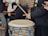 A group of people is gathered for a musical performance, with one individual playing a large drum using drumsticks. Another person is seen with a guitar, while another holds a smartphone. Everyone is wearing warm clothing, indicating a cold environment. The setting is casual and outdoors, with a brick wall visible in the background.
