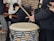 A group of people is gathered for a musical performance, with one individual playing a large drum using drumsticks. Another person is seen with a guitar, while another holds a smartphone. Everyone is wearing warm clothing, indicating a cold environment. The setting is casual and outdoors, with a brick wall visible in the background.