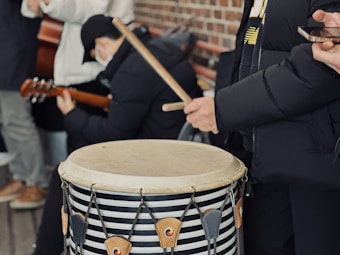 A group of people is gathered for a musical performance, with one individual playing a large drum using drumsticks. Another person is seen with a guitar, while another holds a smartphone. Everyone is wearing warm clothing, indicating a cold environment. The setting is casual and outdoors, with a brick wall visible in the background.