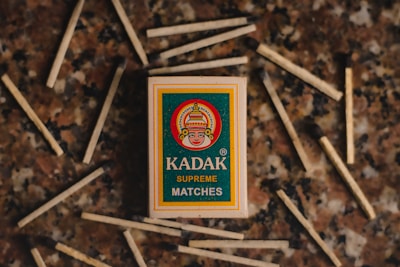 A matchbox labeled 'Kadak Supreme Matches' is surrounded by scattered matchsticks on a textured surface. The matchbox features a stylized image of a face wearing a headdress inside a circular design. The background consists of a marbled pattern in shades of brown, beige, and black.