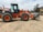 A large orange and black construction vehicle, specifically a Hitachi wheel loader, is parked on a dirt area. The loader has a prominent front bucket used for excavation and construction projects. In the background, there are some trees and another piece of construction equipment. The surrounding area appears to be a construction site with a cloudy sky overhead.