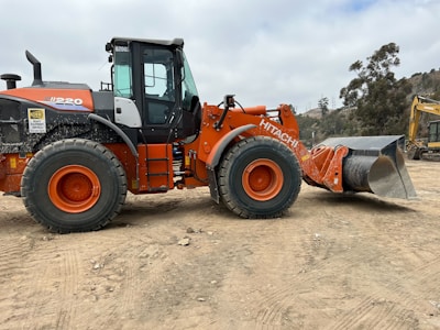 A large orange and black construction vehicle, specifically a Hitachi wheel loader, is parked on a dirt area. The loader has a prominent front bucket used for excavation and construction projects. In the background, there are some trees and another piece of construction equipment. The surrounding area appears to be a construction site with a cloudy sky overhead.