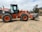 A large orange and black construction vehicle, specifically a Hitachi wheel loader, is parked on a dirt area. The loader has a prominent front bucket used for excavation and construction projects. In the background, there are some trees and another piece of construction equipment. The surrounding area appears to be a construction site with a cloudy sky overhead.