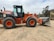 A large orange and black construction vehicle, specifically a Hitachi wheel loader, is parked on a dirt area. The loader has a prominent front bucket used for excavation and construction projects. In the background, there are some trees and another piece of construction equipment. The surrounding area appears to be a construction site with a cloudy sky overhead.