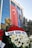 A large glass-fronted building features a prominent Turkish flag and a large portrait of a man. In front is a bust of a person surrounded by vibrant wreaths, composed of red and white flowers. Evergreen trees are visible on the side, contributing to the scenery.