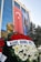 A large glass-fronted building features a prominent Turkish flag and a large portrait of a man. In front is a bust of a person surrounded by vibrant wreaths, composed of red and white flowers. Evergreen trees are visible on the side, contributing to the scenery.