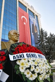 A large glass-fronted building features a prominent Turkish flag and a large portrait of a man. In front is a bust of a person surrounded by vibrant wreaths, composed of red and white flowers. Evergreen trees are visible on the side, contributing to the scenery.