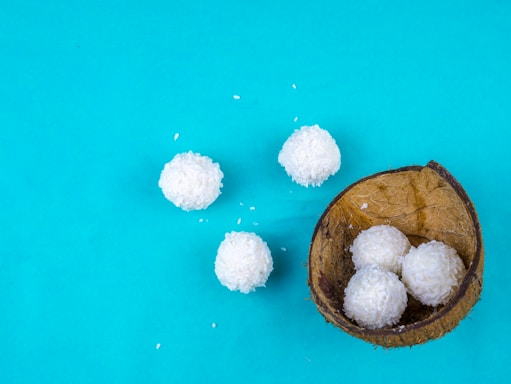 Five white coconut balls are placed on a bright turquoise background. Four of them are inside a half coconut shell, and one is placed beside it. Small coconut flakes are scattered around, adding texture to the image.