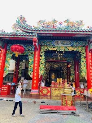 A traditional Chinese temple with ornate red and green decorations, featuring dragon motifs and intricate carvings. Large red columns frame the entrance, which is adorned with golden accents and Chinese characters. Two people are present; one wearing a mask walks by, while another stands near a table. A collection of red and yellow candles is displayed prominently inside the temple.