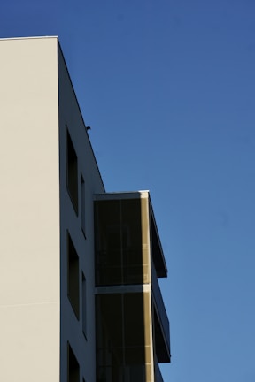 A modern building with a minimalist design, featuring sharp lines and geometric shapes. The facade includes rectangular windows and a section with mesh-like panels. The structure is juxtaposed against a clear blue sky, creating a stark contrast.