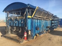 A large, industrial metal structure predominantly painted in blue, possibly used for storage or as a processing unit. It is equipped with numerous pipes, valves, and hatches, with several mechanisms and panels attached. The unit is placed on a sandy terrain, surrounded by an open, clear sky. A red fire extinguisher is positioned near the base.