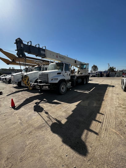 A large white truck crane with the brand name 'Terex' is parked in a construction yard. The crane is aligned with several other construction vehicles, and a bright blue sky is visible in the background. Shadows from the crane are cast prominently on the ground.