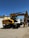 A large yellow CAT M322 excavator stands on a concrete surface outside a building under a clear blue sky. The excavator has an articulated arm and large wheels, and is positioned near a white SUV and a person holding papers. The building has a roll-up garage door.