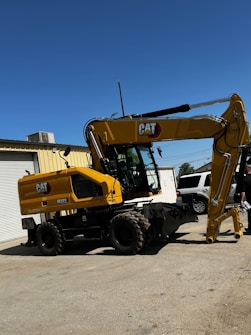 A large yellow CAT M322 excavator stands on a concrete surface outside a building under a clear blue sky. The excavator has an articulated arm and large wheels, and is positioned near a white SUV and a person holding papers. The building has a roll-up garage door.