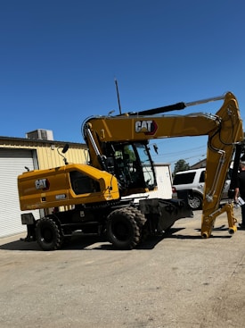 A large yellow CAT M322 excavator stands on a concrete surface outside a building under a clear blue sky. The excavator has an articulated arm and large wheels, and is positioned near a white SUV and a person holding papers. The building has a roll-up garage door.