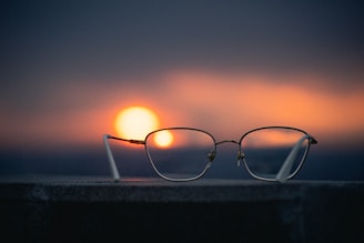 A pair of eyeglasses rests on a concrete surface with a blurred background featuring a vibrant sunset. The sun appears low on the horizon, casting warm orange and yellow hues across the sky, which gradually darkens to a deep blue.
