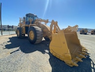 The image features a large, yellow construction vehicle equipped with massive wheels and a front bucket, parked on a gravel surface. The backdrop includes other similar machinery and industrial buildings under a clear blue sky.