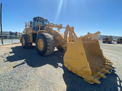 The image features a large, yellow construction vehicle equipped with massive wheels and a front bucket, parked on a gravel surface. The backdrop includes other similar machinery and industrial buildings under a clear blue sky.