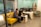 Two people are collaborating in a modern office space. They are seated on a vibrant yellow couch, closely looking at a laptop on the small, matching coffee table. Another person is seated at a white desk with multiple computer monitors. The room has a light teal accent wall and modern furniture.