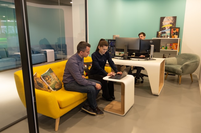 Two people are collaborating in a modern office space. They are seated on a vibrant yellow couch, closely looking at a laptop on the small, matching coffee table. Another person is seated at a white desk with multiple computer monitors. The room has a light teal accent wall and modern furniture.