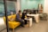 Two people are collaborating in a modern office space. They are seated on a vibrant yellow couch, closely looking at a laptop on the small, matching coffee table. Another person is seated at a white desk with multiple computer monitors. The room has a light teal accent wall and modern furniture.