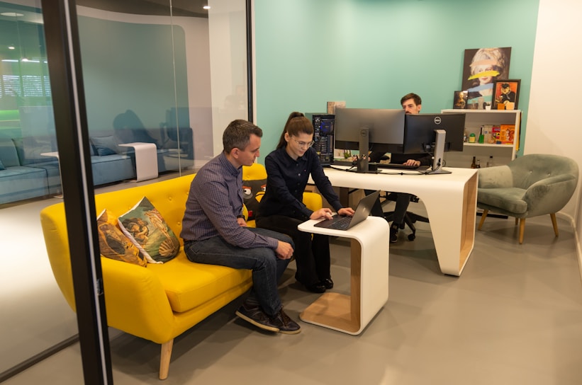 Two people are collaborating in a modern office space. They are seated on a vibrant yellow couch, closely looking at a laptop on the small, matching coffee table. Another person is seated at a white desk with multiple computer monitors. The room has a light teal accent wall and modern furniture.