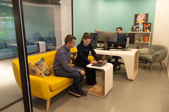 Two people are collaborating in a modern office space. They are seated on a vibrant yellow couch, closely looking at a laptop on the small, matching coffee table. Another person is seated at a white desk with multiple computer monitors. The room has a light teal accent wall and modern furniture.