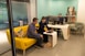Two people are collaborating in a modern office space. They are seated on a vibrant yellow couch, closely looking at a laptop on the small, matching coffee table. Another person is seated at a white desk with multiple computer monitors. The room has a light teal accent wall and modern furniture.