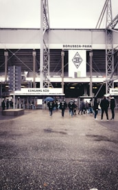 A group of people walking towards the entrance of a stadium with the sign 'Borussia-Park' prominently displayed. The weather appears cloudy, and some individuals are carrying umbrellas. The ground is wet, suggesting recent rain.