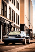 A vintage Jaguar car with a '95-D-49694' license plate is parked on the side of a street lined with tall buildings. The buildings have a classical architectural style with rectangular windows and ornate details around the entrances. The street has a slightly worn look, and the sky appears to be clear.