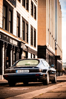 A vintage Jaguar car with a '95-D-49694' license plate is parked on the side of a street lined with tall buildings. The buildings have a classical architectural style with rectangular windows and ornate details around the entrances. The street has a slightly worn look, and the sky appears to be clear.
