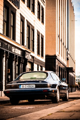 A vintage Jaguar car with a '95-D-49694' license plate is parked on the side of a street lined with tall buildings. The buildings have a classical architectural style with rectangular windows and ornate details around the entrances. The street has a slightly worn look, and the sky appears to be clear.