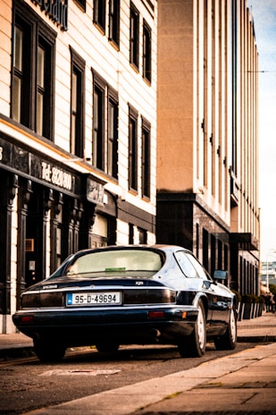 A vintage Jaguar car with a '95-D-49694' license plate is parked on the side of a street lined with tall buildings. The buildings have a classical architectural style with rectangular windows and ornate details around the entrances. The street has a slightly worn look, and the sky appears to be clear.