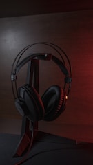 A pair of black over-ear headphones is placed on a sleek metal stand. The background has a muted, dark tone with a subtle glow of red light adding a dramatic effect. The headphones have a cushioned design with visible branding on the earpiece.