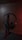 A pair of black over-ear headphones is placed on a sleek metal stand. The background has a muted, dark tone with a subtle glow of red light adding a dramatic effect. The headphones have a cushioned design with visible branding on the earpiece.