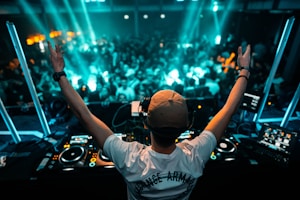 A DJ stands at a mixing console with arms raised, facing a crowded nightclub filled with people dancing under blue lights. The equipment is illuminated, and visual screens are positioned around the DJ booth, contributing to the lively atmosphere.