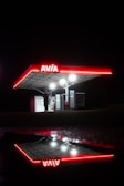A brightly illuminated gas station canopy with the red Avia logo is set against a dark night background. The structure is reflected on a wet surface, creating a mirror image. Underneath the canopy, a person stands near the gas pumps, partially silhouetted by the lights.