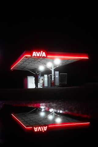 A brightly illuminated gas station canopy with the red Avia logo is set against a dark night background. The structure is reflected on a wet surface, creating a mirror image. Underneath the canopy, a person stands near the gas pumps, partially silhouetted by the lights.