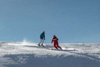 Two skiers are descending a snow-covered slope under a clear blue sky. One person is dressed in a turquoise jacket with white pants and the other in a red ski suit. Both are wearing helmets and goggles, skillfully navigating through the snow while leaving trails behind them.