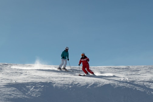 Two skiers are descending a snow-covered slope under a clear blue sky. One person is dressed in a turquoise jacket with white pants and the other in a red ski suit. Both are wearing helmets and goggles, skillfully navigating through the snow while leaving trails behind them.