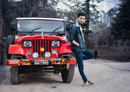 A man in a denim jacket and jeans casually leans against a bright red off-road vehicle with a Pakistan tourism sticker on the front. The scene is set on a dirt path surrounded by trees, and another person is visible in the distant background.