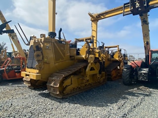 A lineup of heavy construction machinery, including large bulldozers and other vehicles, set on a gravel surface. The equipment features prominent treads and metal arms, indicating their use for construction and heavy-duty tasks.